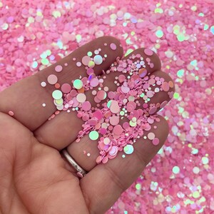 May include: A close-up of a hand holding pink and iridescent glitter in various sizes. The glitter is scattered on a surface and the hand is holding a small amount of the glitter.