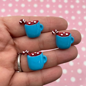 May include: Three blue ceramic mugs filled with red hot chocolate and white marshmallows. Each mug has a red and white candy cane sticking out of the top.