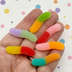 May include: A handful of colorful gummy worms. Each worm is a different color combination, including green and yellow, red and yellow, pink and orange, and blue and orange. The candies are on a white background with colorful polka dots.
