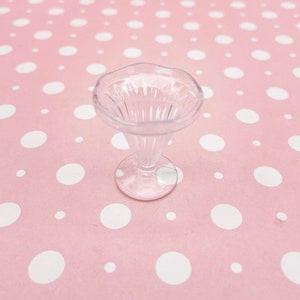 May include: A clear plastic sundae dish with a fluted rim and a pedestal base. The dish is empty and sits on a pink background with white polka dots.