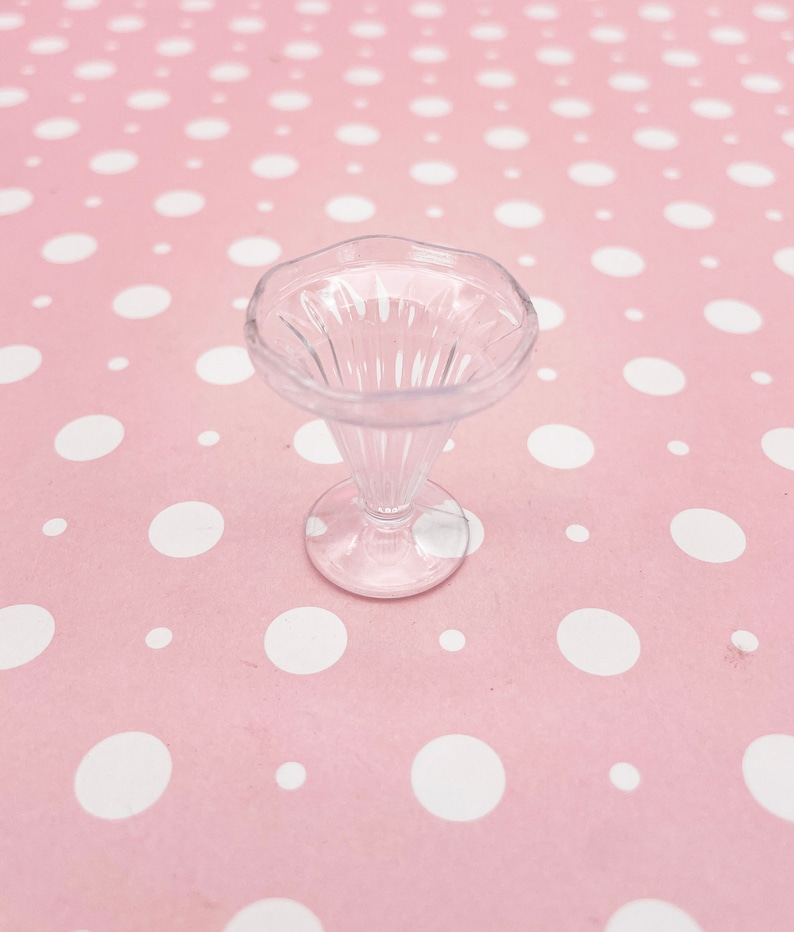 May include: A clear plastic sundae dish with a fluted rim and a pedestal base. The dish is empty and sits on a pink background with white polka dots.
