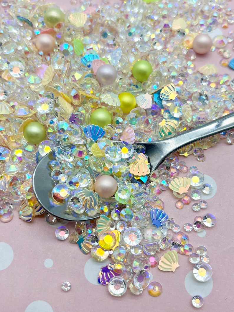 May include: A mix of iridescent clear and pastel-colored glitter, seashells, and pearls. The glitter is in various shapes and sizes, including round, square, and star-shaped. The seashells are in various colors, including white, pink, and blue. The pearls are in various sizes and colors, including white, pink, and green.
