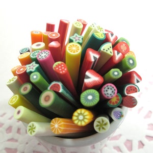 May include: A collection of colorful polymer clay sticks with various fruit designs, including watermelon, kiwi, pineapple, and strawberries. The sticks are arranged in a white cup.