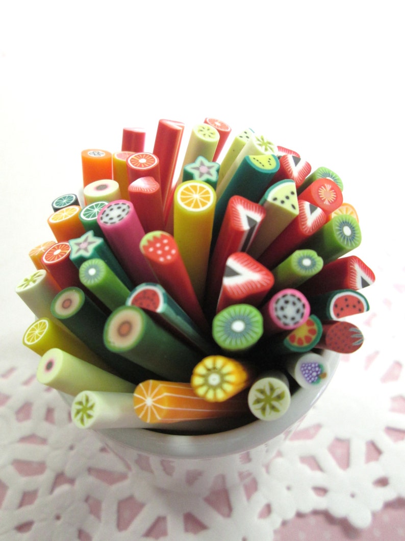 May include: A collection of colorful polymer clay sticks with various fruit designs, including watermelon, kiwi, pineapple, and strawberries. The sticks are arranged in a white cup.
