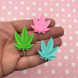 May include: Three resin cannabis leaf charms in pink, green, and light blue. The charms are about 2 inches in size and have a textured surface.