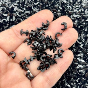 May include: A collection of small, black plastic clips, some held in a hand. The clips have a curved, C-shaped design with a small post on one side. The background is filled with more of the same clips.