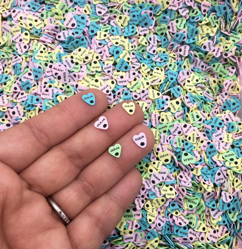 May include: A hand holding three small heart-shaped charms with the word "OUIJA" printed on them. The charms are green, pink, and blue. The background is a large pile of similar charms in various colors.