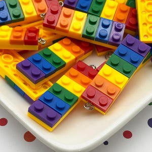 May include: A collection of colorful, rainbow-themed brick pendants. Each pendant is made of small, rectangular blocks in a spectrum of colors, including red, orange, yellow, green, blue, and purple. The pendants are on a white plate with a polka dot background.