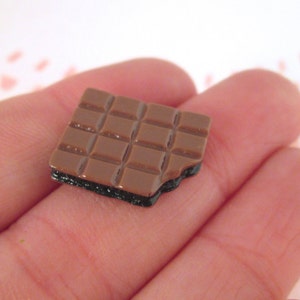 Puede incluir: Una pequeña barra de chocolate con un envoltorio marrón y negro. La barra está dividida en cuadrados y tiene una forma ligeramente irregular.
