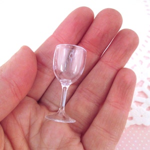 May include: A clear plastic wine glass, approximately 5 cm tall, is held in a person's hand. The glass is small and delicate, perfect for dollhouses or miniature settings.