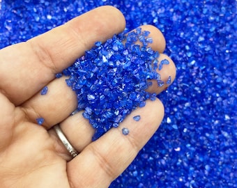 May include: A handful of vibrant blue crushed glass, with a sparkling, reflective quality. The glass pieces are irregular in shape and size, creating a textured appearance. The background is filled with more of the same blue glass.