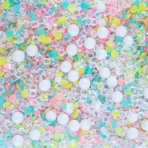 Könnte beinhalten: Eine Mischung aus pastellfarbenen Streuseln, darunter weiß, rosa, gelb, blau und grün. Die Streusel haben verschiedene Formen, darunter runde, quadratische und sternförmige.