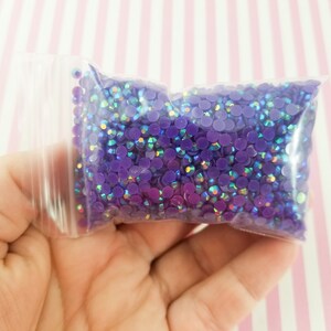 May include: A clear plastic bag filled with small, round, purple rhinestones. The rhinestones have an iridescent sheen, reflecting multiple colors. The bag is held in a hand, with a pink and white striped background.