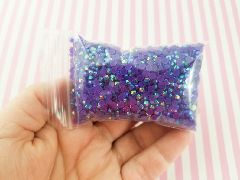 May include: A clear plastic bag filled with small, round, purple rhinestones. The rhinestones have an iridescent sheen, reflecting multiple colors. The bag is held in a hand, with a pink and white striped background.