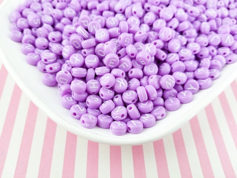 Peut inclure: Un grand tas de perles de lettres violettes avec des lettres blanches. Les perles sont sur une assiette blanche avec un fond ray&eacute; rose et blanc.