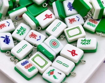 May include: A collection of small, rectangular Mahjong tile charms. The charms are white with green edges and feature various symbols and characters in red, blue, and green. Each charm has a silver-colored loop.