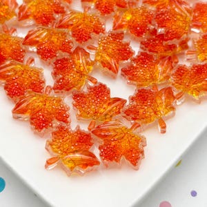 May include: A collection of orange and yellow maple leaf-shaped embellishments. The translucent leaves have a textured, speckled appearance and are arranged on a white surface. These decorative elements are ideal for fall-themed crafts.
