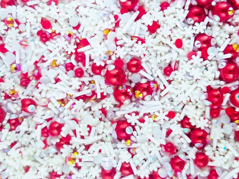 May include: A close-up of a mixture of red and white sprinkles, including snowflake shapes, small round sprinkles, and long, thin sprinkles.