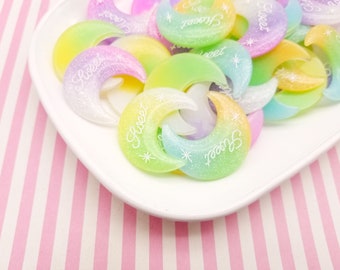 May include: A collection of pastel-colored crescent moon shaped resin charms with the word "Sweet" written on them.
