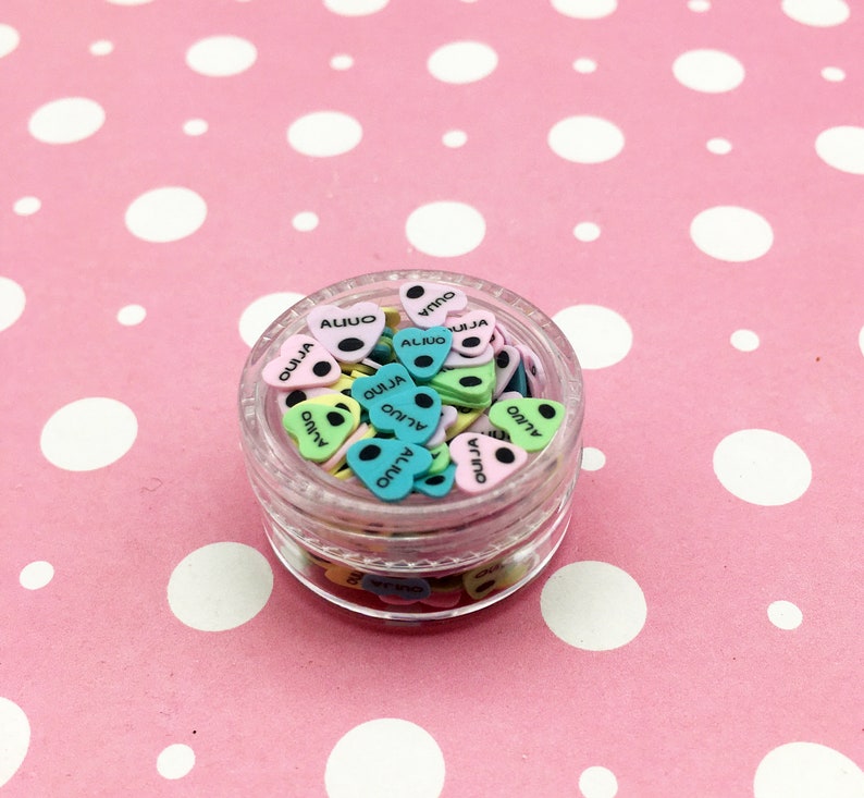 May include: A clear plastic jar filled with colorful heart-shaped charms. The charms are decorated with black dots and the words "Ouija", "Virgo", and "Aliuo".