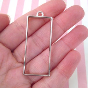 Puede incluir: Pendiente de metal plateado con un marco rectangular. El marco está abierto por todos lados y tiene un bucle en la parte superior para sujetar una cadena o un cordón.