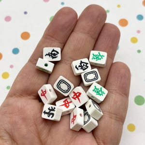 May include: A collection of small, white Mahjong tiles with black, red, and green symbols. The tiles are held in a hand, showcasing the variety of characters and designs used in the game. The background is a colorful pattern of polka dots.