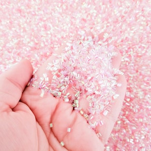 May include: A hand holding a pile of iridescent pink and white glass beads. The beads are small and have a square shape.