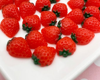 May include: A collection of red and green plastic strawberry shaped charms. The strawberries are arranged on a white plate.