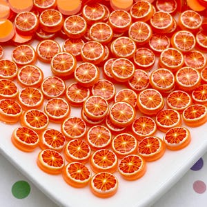 Puede incluir: Una colección de cabujones de resina naranja y roja, cada uno con un diseño detallado de rodaja de naranja. Los cabujones están dispuestos sobre una superficie blanca, mostrando sus colores vibrantes y patrones intrincados. Ideales para manualidades y joyería.