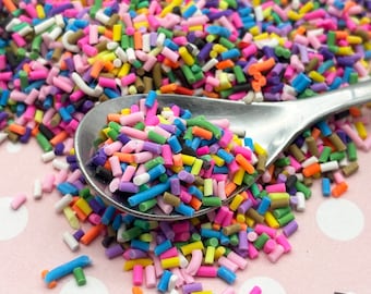 May include: A spoon filled with colorful sprinkles, a mix of pink, blue, green, yellow, and white. The sprinkles are scattered on a pink surface with white polka dots.