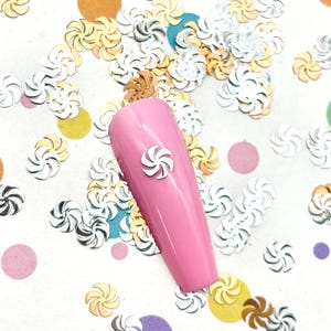 May include: A glossy pink artificial nail tip featuring a small, white, spiral-shaped embellishment centered on its surface, surrounded by a scattered collection of metallic gold, silver, and white swirl-shaped confetti pieces.