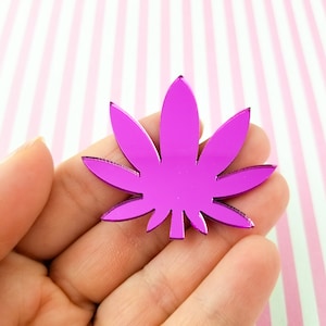 May include: A shiny, magenta-colored cannabis leaf-shaped charm. The charm is held in a hand against a background of pink and white stripes. The charm has a reflective surface.