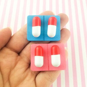 May include: Two sets of pill-shaped earrings, one set on a blue backing and one set on a pink backing. Each set has two earrings, each with a red top and a white bottom.