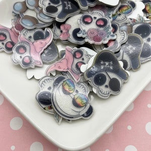 Può includere: Una collezione di ciondoli a forma di teschio in resina colorati e scintillanti con vari design, tra cui un teschio con un cappello da pirata, un teschio con fiori e un teschio con occhiali da sole. I ciondoli sono su un piatto bianco con uno sfondo a pois rosa.