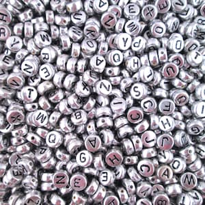 Puede incluir: Una imagen de primer plano de un mont&oacute;n grande de cuentas de letras plateadas. Las cuentas son redondas y tienen un agujero en el centro. Cada cuenta tiene una sola letra impresa en negro.