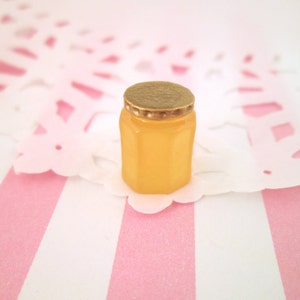 May include: A small, clear glass jar with a gold lid. The jar is filled with a yellow liquid, possibly honey. The jar is sitting on a white and pink striped surface.