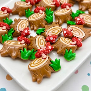 May include: A collection of miniature resin forest-themed embellishments. The set includes brown tree stumps with light brown spiral details, red mushrooms with white spots, and green grass accents. These decorative pieces are arranged on a white surface.