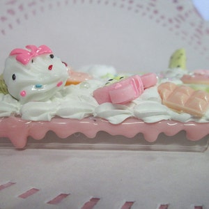 May include: A decorative phone case featuring a whimsical design. The case is adorned with white frosting, pink dripping accents, and miniature candy decorations, including an ice cream cone, a popsicle, and chocolate bars. The overall aesthetic is playful and sweet.