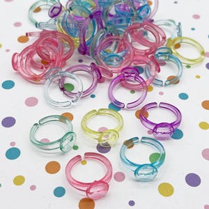 May include: A collection of colorful plastic rings in various shades of pink, purple, blue, green, and yellow. Each ring features a clear, oval-shaped gem. The rings are scattered on a white surface with colorful polka dots.