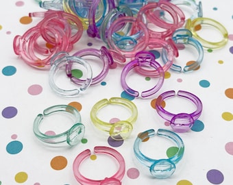 May include: A collection of colorful plastic rings in various shades of pink, purple, blue, green, and yellow. Each ring features a clear, oval-shaped gem. The rings are scattered on a white surface with colorful polka dots.