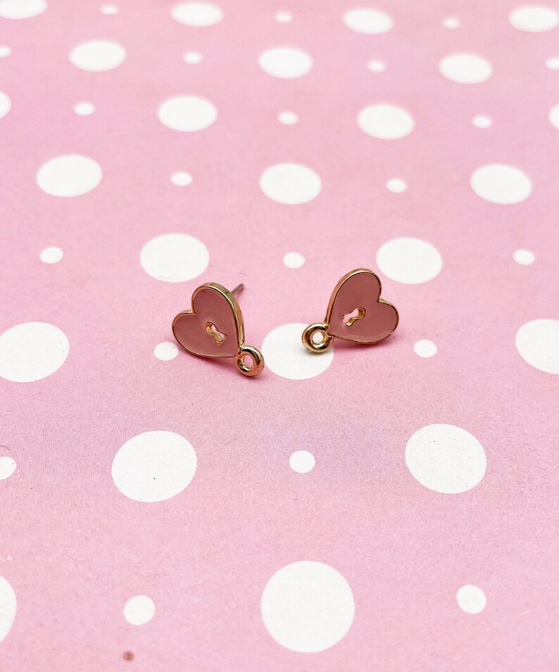 May include: A pair of heart-shaped stud earrings. Each earring features a light pink enamel heart with a keyhole design and a gold-colored metal border and post. The earrings are displayed on a pink background with white polka dots.