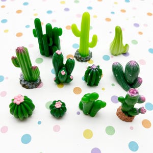 May include: A collection of eleven miniature cactus figurines in various shades of green, some featuring small pink flower accents and textured brown bases, arranged in a group on a surface with a colorful polka dot pattern.