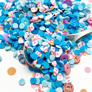 Op de afbeelding: Een verscheidenheid aan blauwe, roze en witte harsstrooisels in een lepel. De strooisels zijn gevormd als donuts, harten en flessen.
