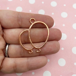 May include: Rose gold-colored metal balloon charm with two connected balloon shapes. The charm has a loop at the top for hanging and small loops at the bottom of each balloon. The background is a light pink color with white polka dots.