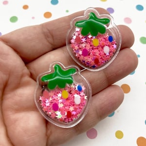 Op de afbeelding: Twee heldere aardbeivormige bedels gevuld met roze glitter en kleurrijke confetti. De bedels hebben groene toppen en worden in een hand gehouden. De achtergrond is wit met kleurrijke stippen.