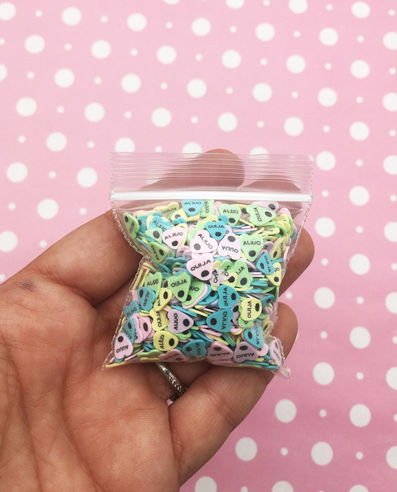 May include: A clear plastic bag filled with colorful heart-shaped sprinkles. The sprinkles are decorated with the words "OUIJA" and "ALINO" in black lettering.
