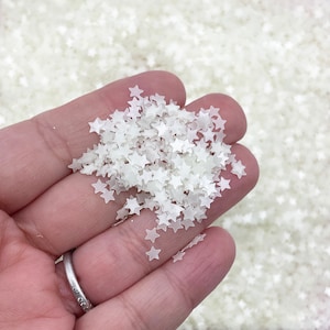 Pode incluir: Uma mão segura uma coleção de pequenas decorações translúcidas em forma de estrela. As estrelas são de um tom branco-sujo pálido e parecem delicadas. O fundo é preenchido com mais estrelas iguais, criando um efeito texturizado.