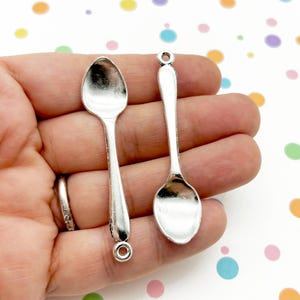 Puede incluir: Dos pequeños colgantes de cuchara plateados. Cada cuchara tiene un cuenco redondeado y un mango largo con un lazo en el extremo. Las cucharas se muestran sobre un fondo de lunares de colores.