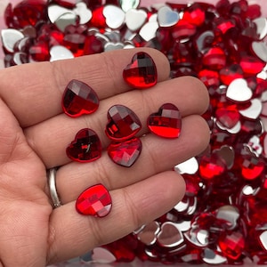 May include: A hand holding a variety of red heart-shaped rhinestones. The rhinestones are faceted and have a shiny finish.