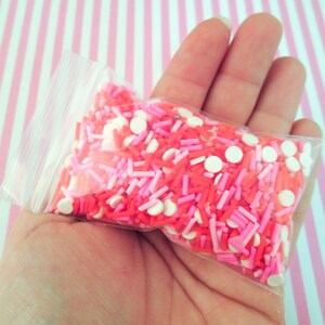 May include: A clear plastic bag filled with pink, red, and white sprinkles. The sprinkles are a mix of small round sprinkles and long, thin sprinkles.